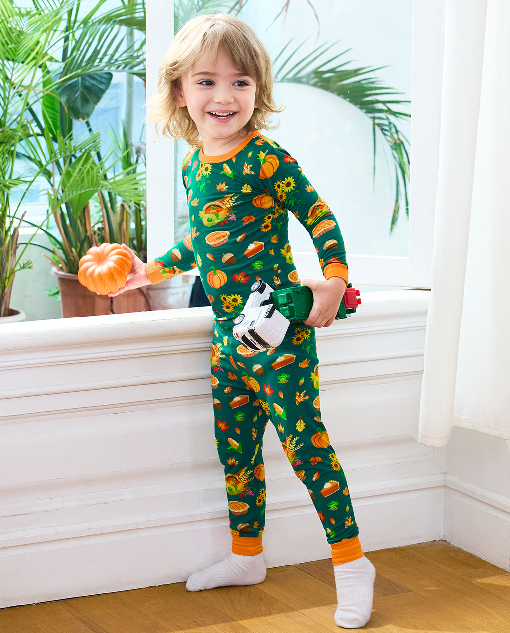 Child wearing Harvest Feast long two-piece pajamas beside a window while holding toy trucks and a pumpkin.