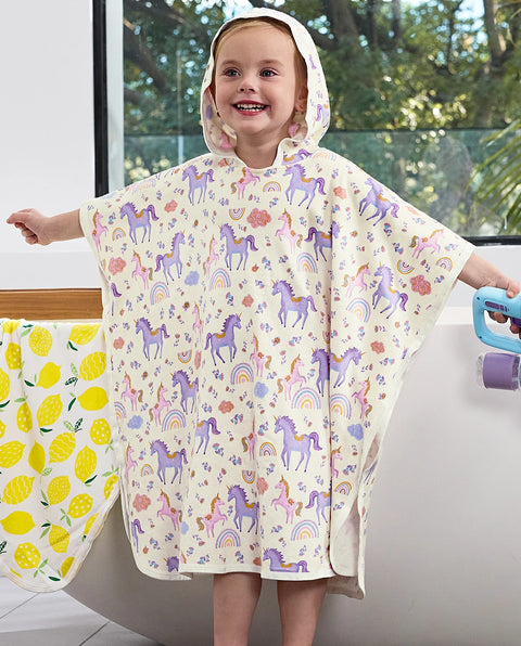 A young girl smiling while wearing a cream hooded poncho with a colorful print of purple unicorns, rainbows, and flowers. She is standing in front of a bathtub with a yellow lemon-patterned towel on the left.