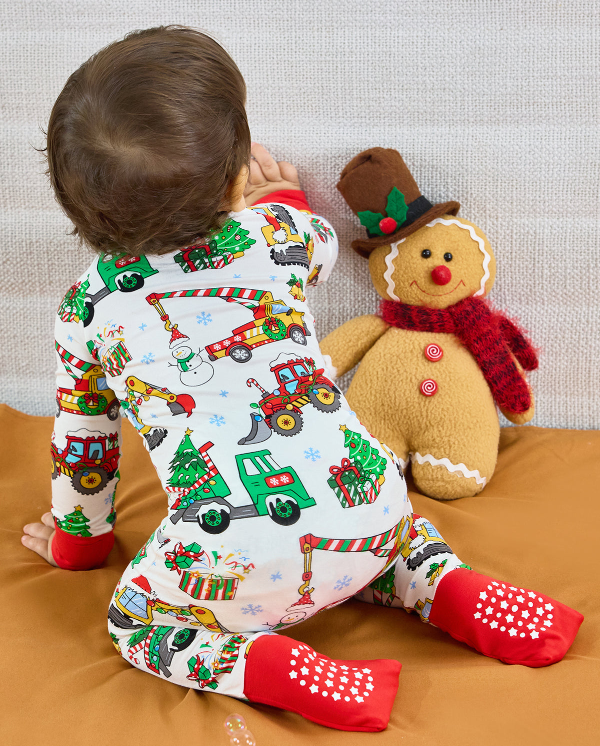 Rear view of a child kneeling in the Merry Truckmas Bamboo Zippy, showing red foot cuffs with white grip dots beside a gingerbread toy.