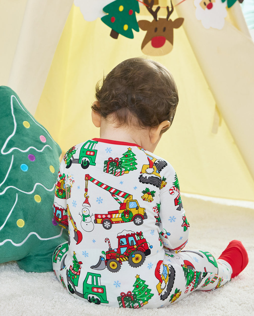 Child seated beside a Christmas tree cushion in the Merry Truckmas Bamboo Zippy, showing the printed back and red foot cuff.