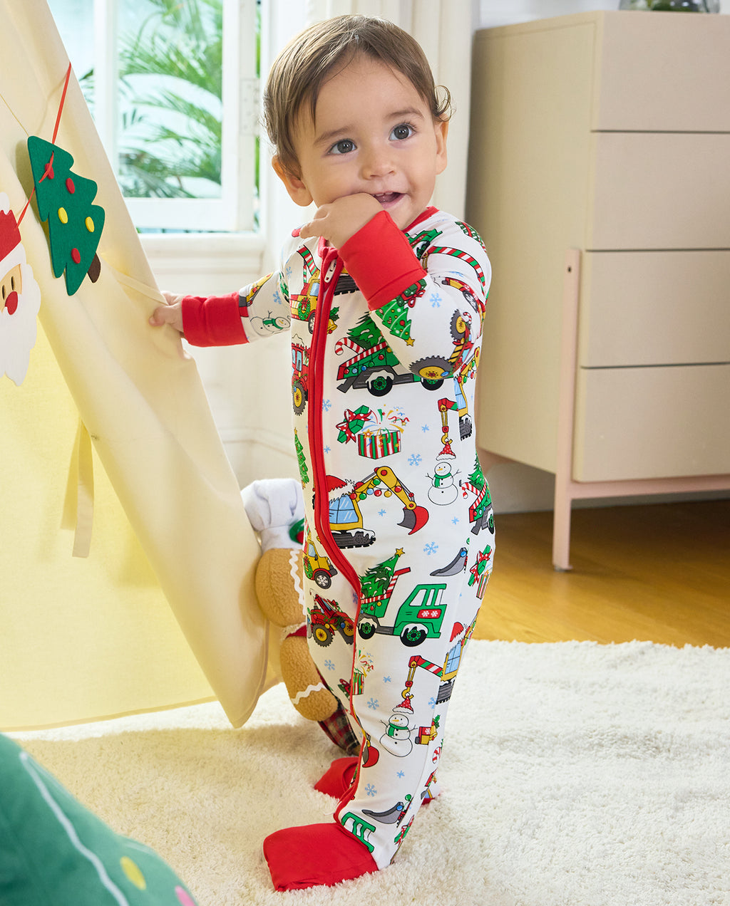 Child standing beside a festive curtain in the Merry Truckmas Bamboo Zippy, showing the full front, red zipper and covered feet.