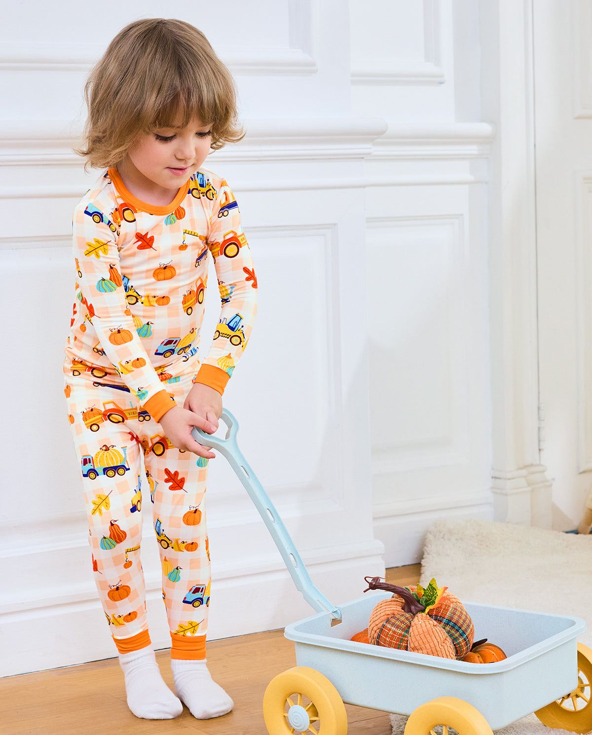 Child wearing Pumpkin Tractors long two-piece pajamas beside a toy wagon with pumpkins.